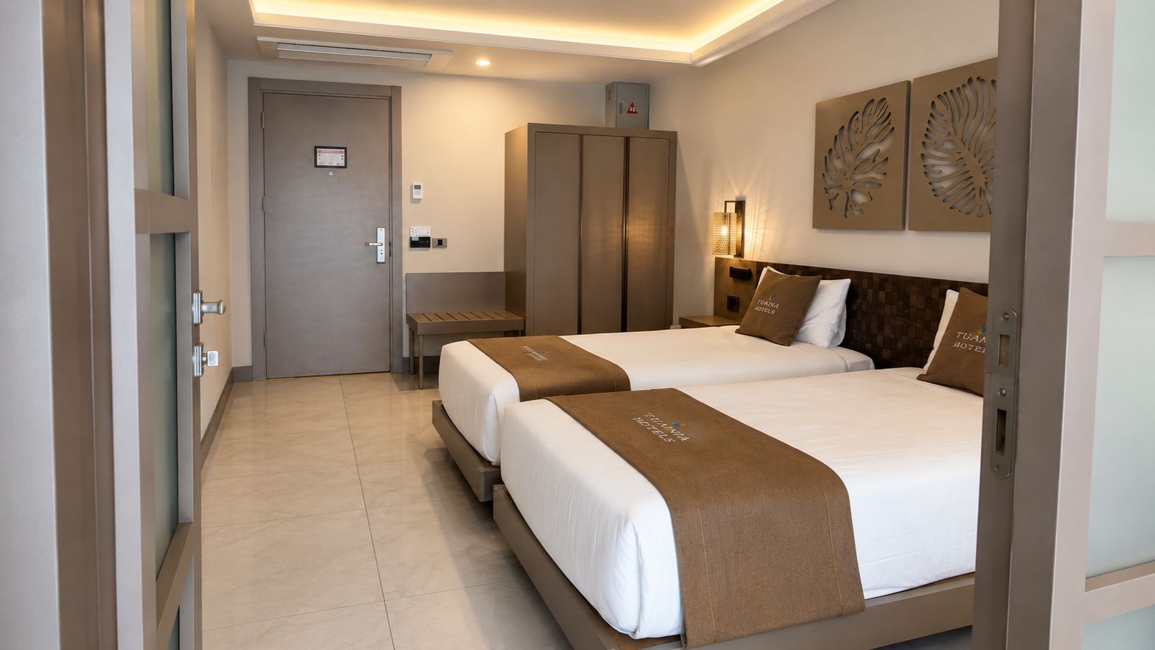 A hotel room with two beds, a wardrobe, a desk, and a wall-mounted light fixture.