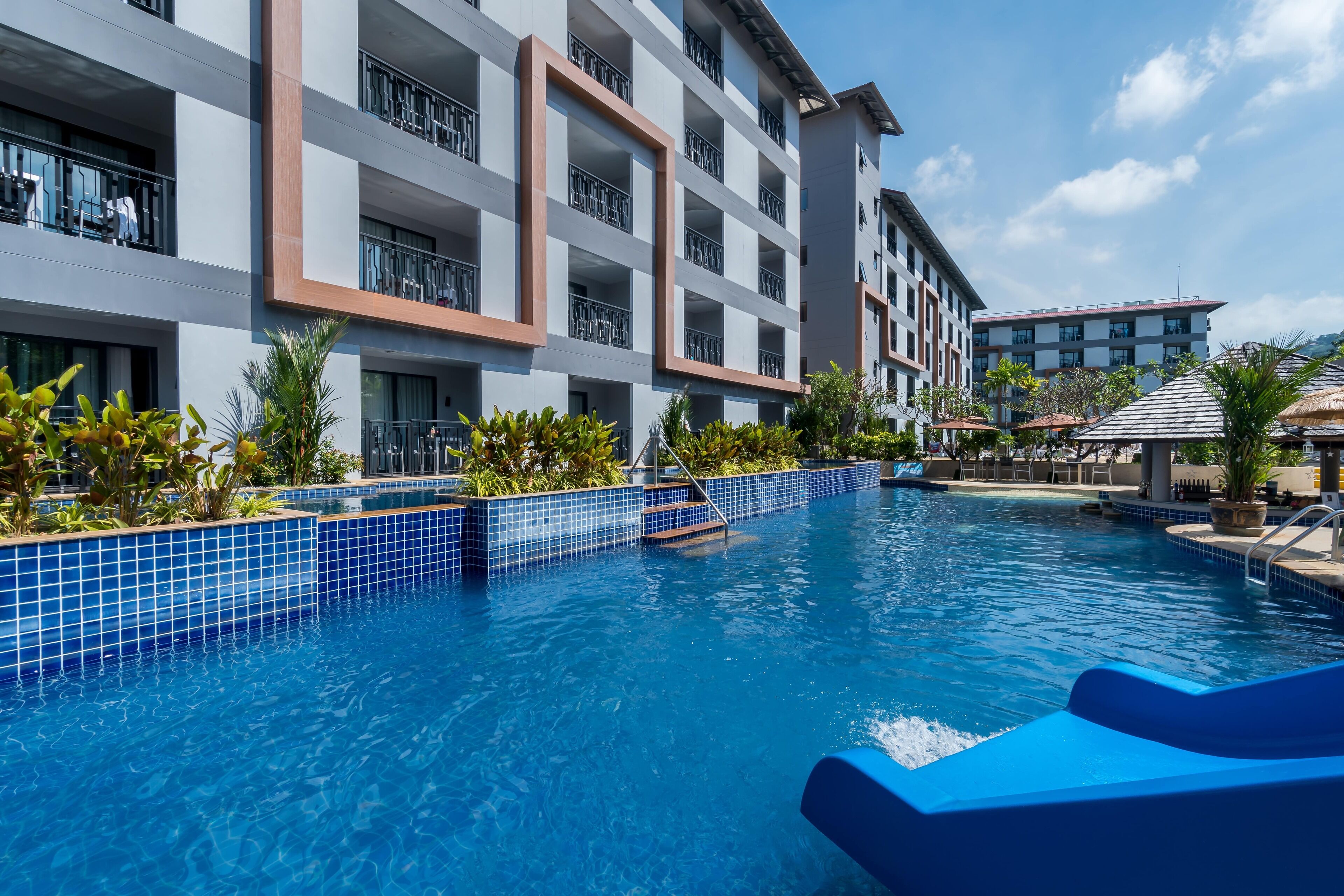 Una piscina con bordes de azulejos azules, rodeada por un edificio de hotel de varios pisos con balcones.