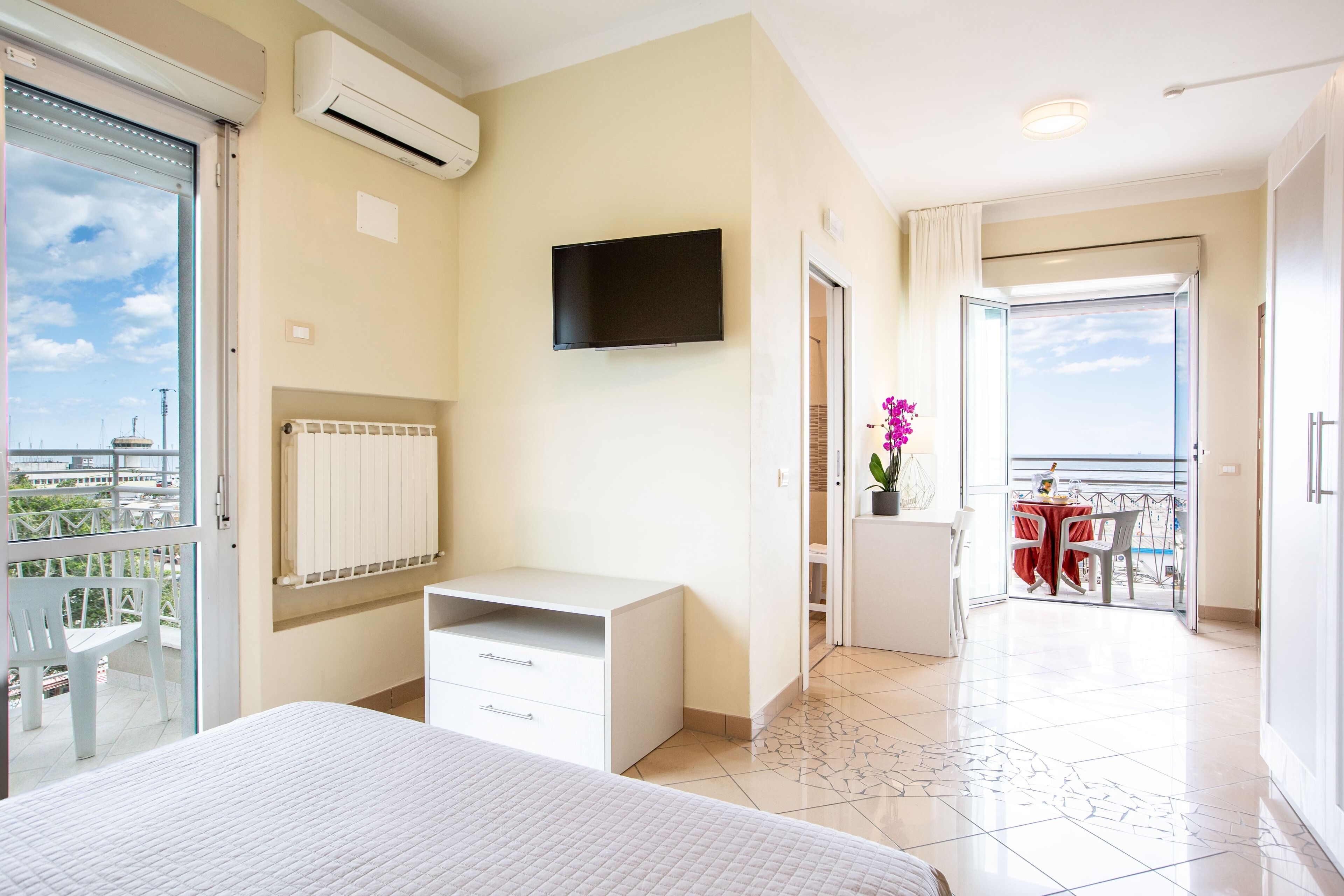 A hotel room with a bed, a TV, a radiator, a balcony with a table and chairs, and a view of the city.