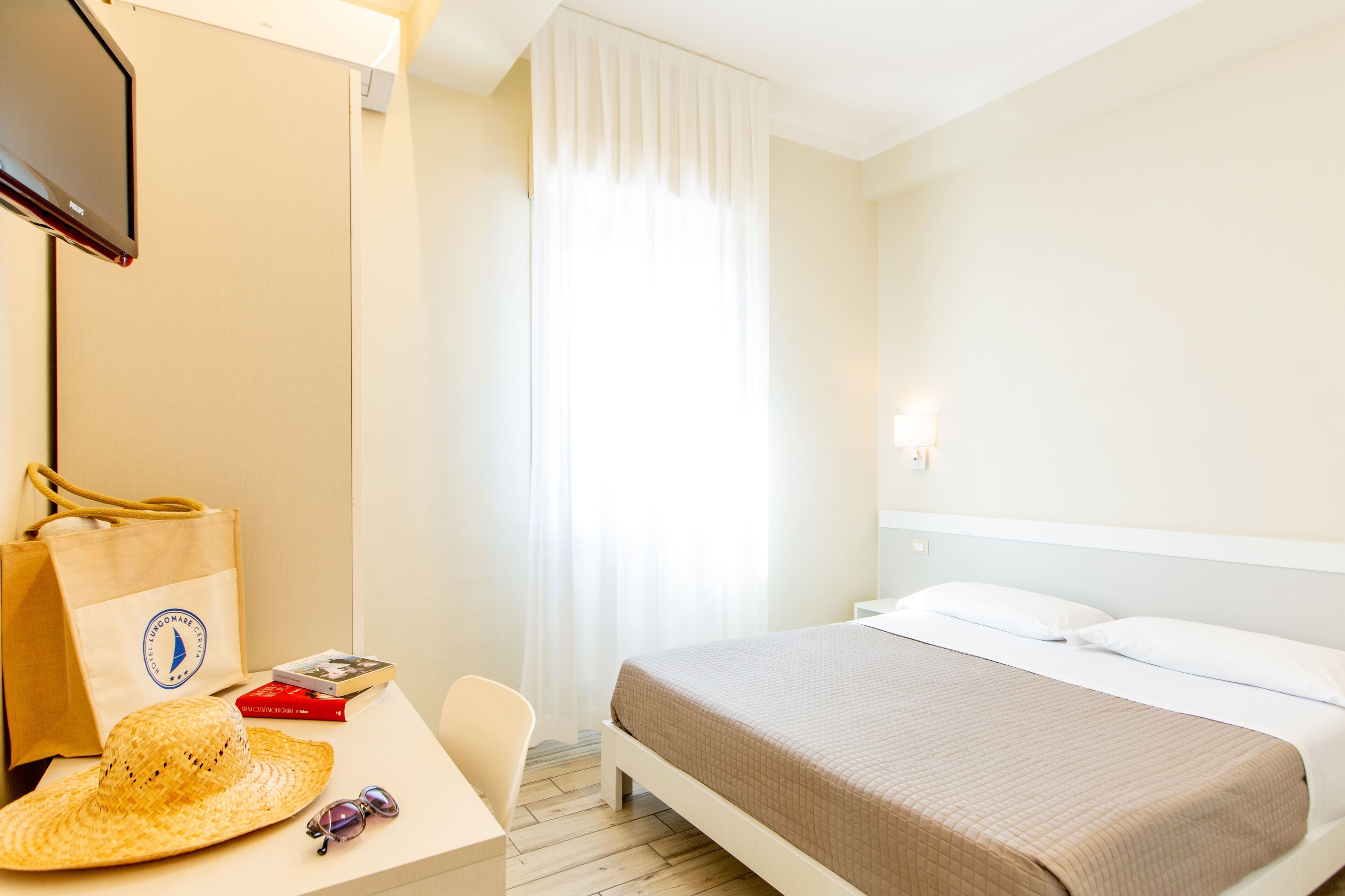 Hotellihuone, jossa on sänky, työpöytä tuolilla, televisio, ikkuna verhoilla ja rantakassi.