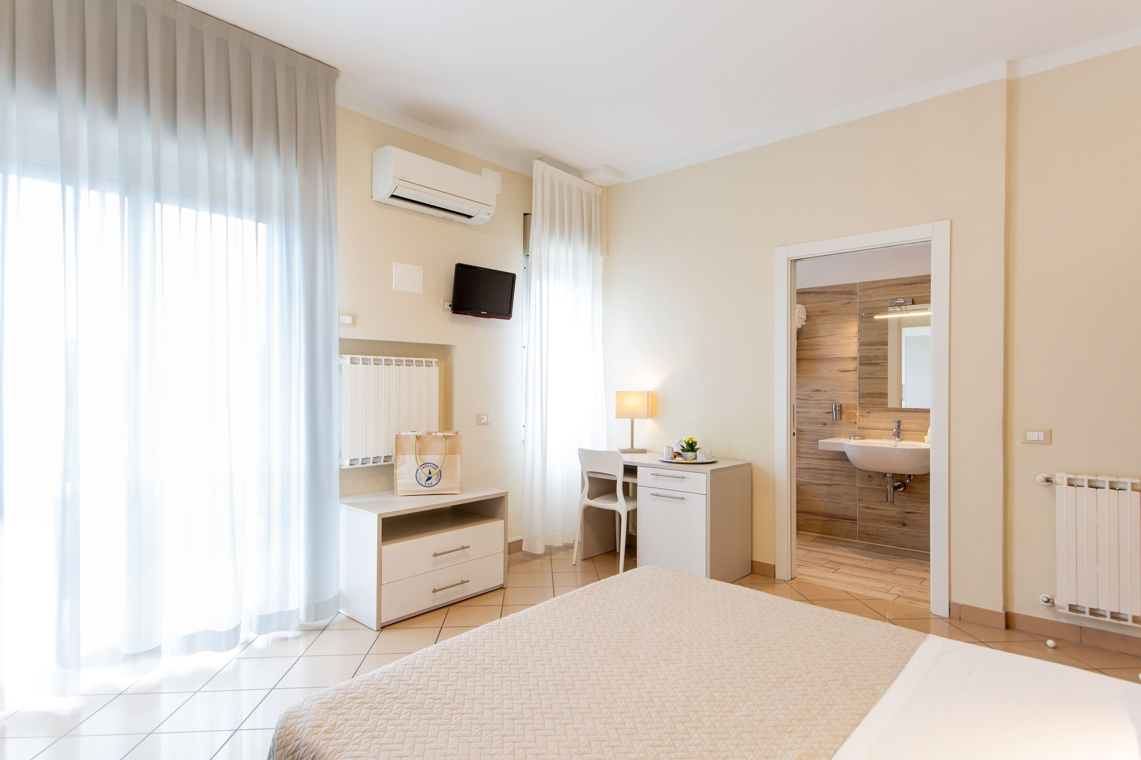 A hotel room with a bed, a desk, a chair, a TV, a bathroom with a sink, and a window with curtains.
