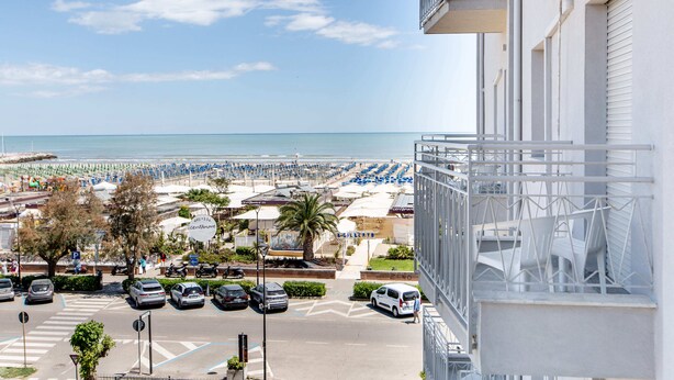 Vista de un estacionamiento frente a la playa con coches, un paso de peatones y un edificio con balcón que da al mar.