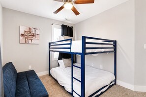 Une chambre avec un lit superposé, un ventilateur de plafond, une fenêtre avec des rideaux, un tableau décoratif au mur et un canapé bleu foncé.