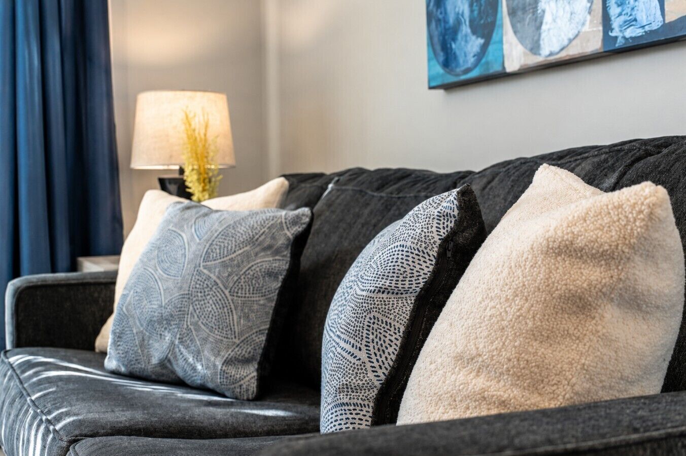 Ein dunkelgraues Sofa mit gemusterten Kissen und eine Lampe mit einer gelben Blumenanordnung auf einem Beistelltisch.