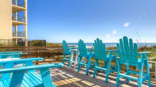 A wooden deck with turquoise Adirondack chairs and a building with balconies.