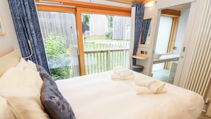 A neatly made bed with white linens and two towels, a wooden headboard, and a view of a wooden deck outside. - Lodge 4 (Wareham)