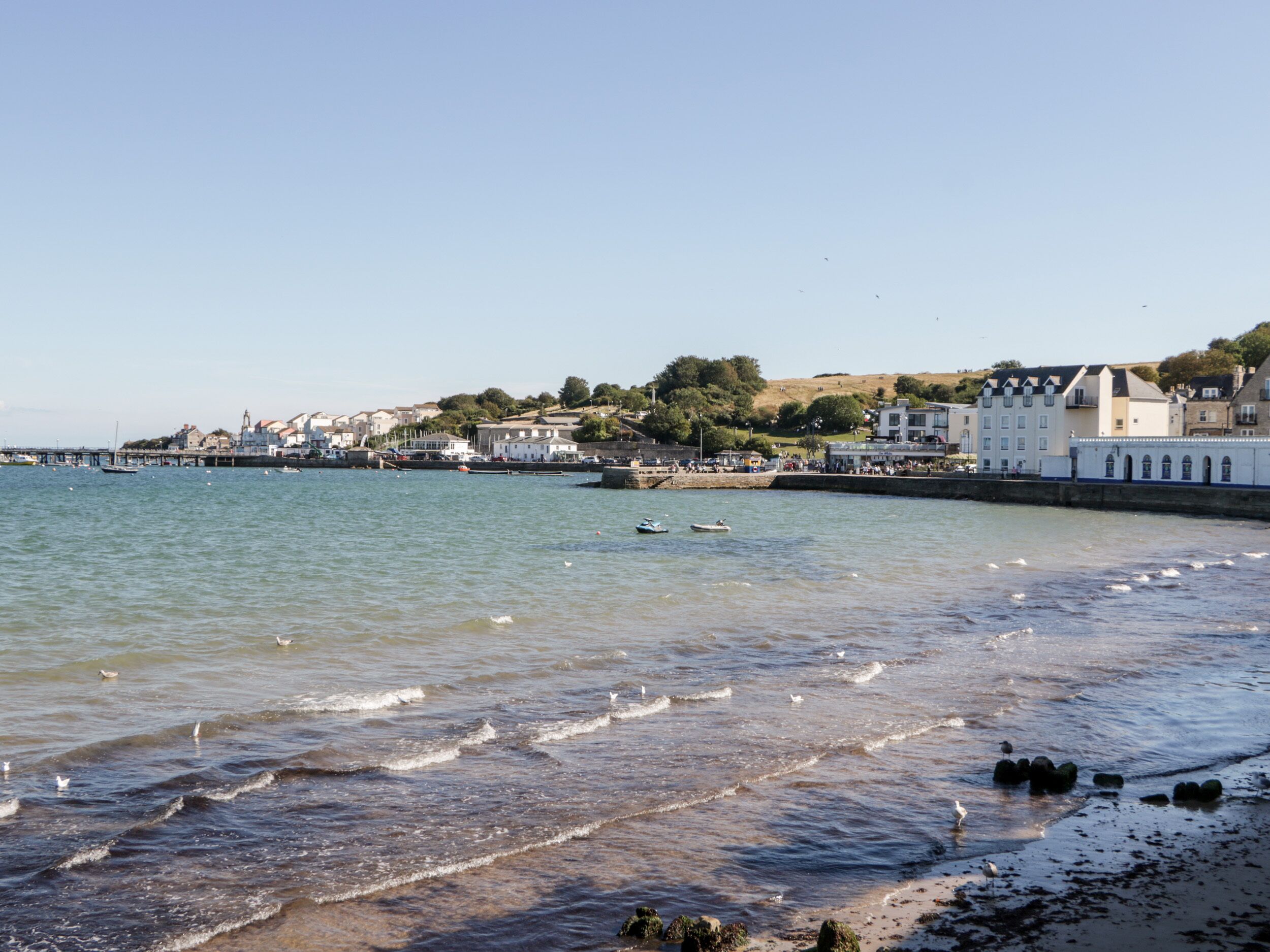 A coastal town with a pier extending into calm waters, buildings along the shore, and a clear sky.