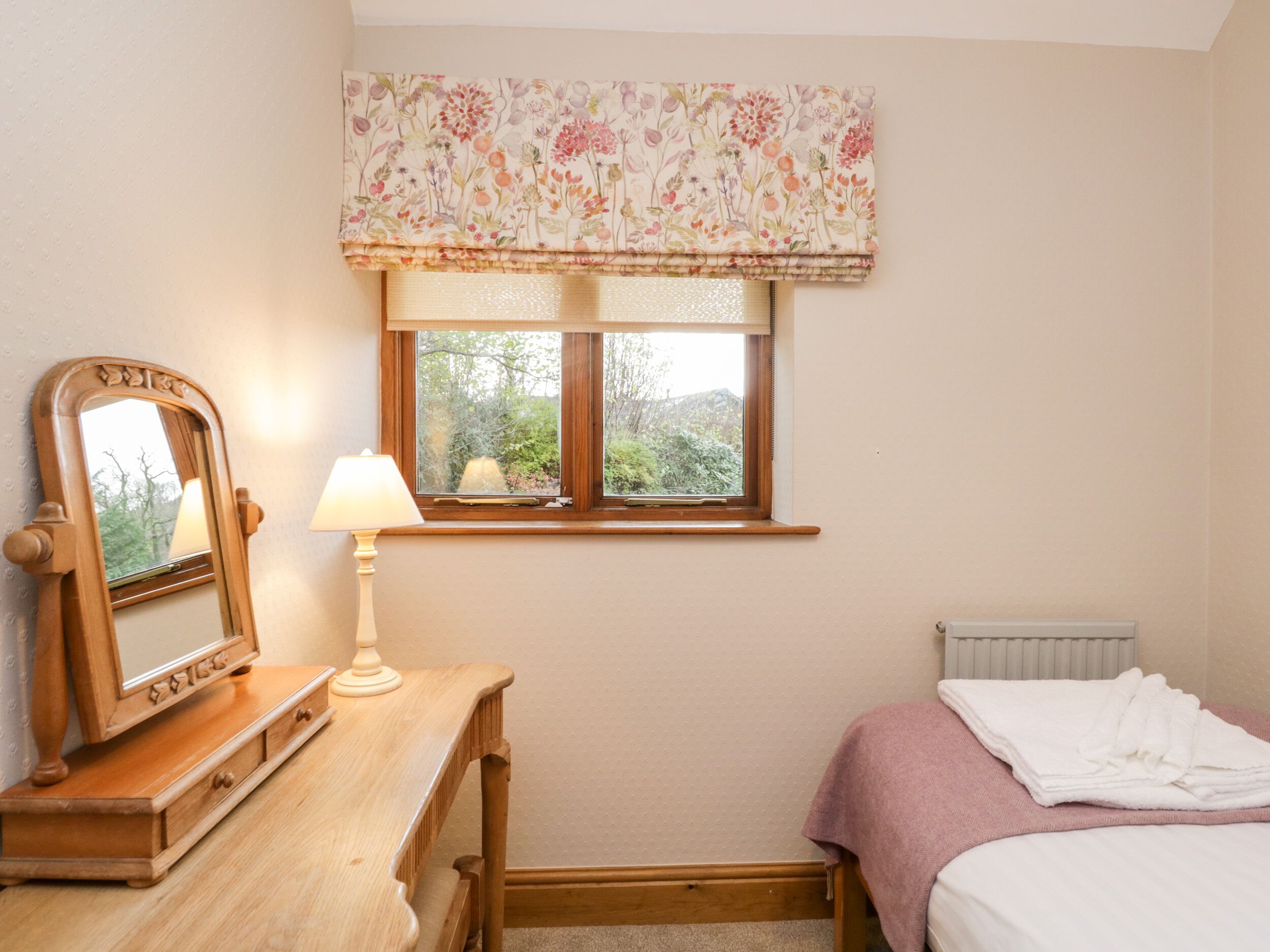 A bedroom with a wooden dresser, a mirror, a bedside lamp, a window with a floral valance, and a bed with a pink blanket.