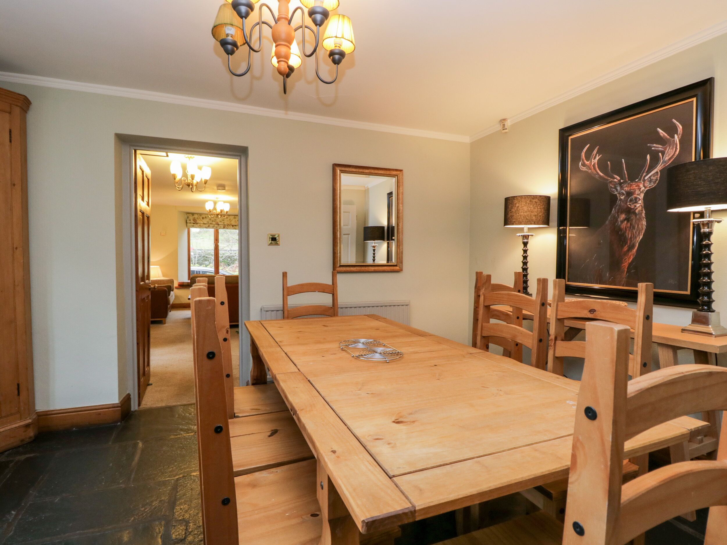 Comedor con mesa y sillas de madera, una araña, un espejo y un cuadro de un ciervo enmarcado.