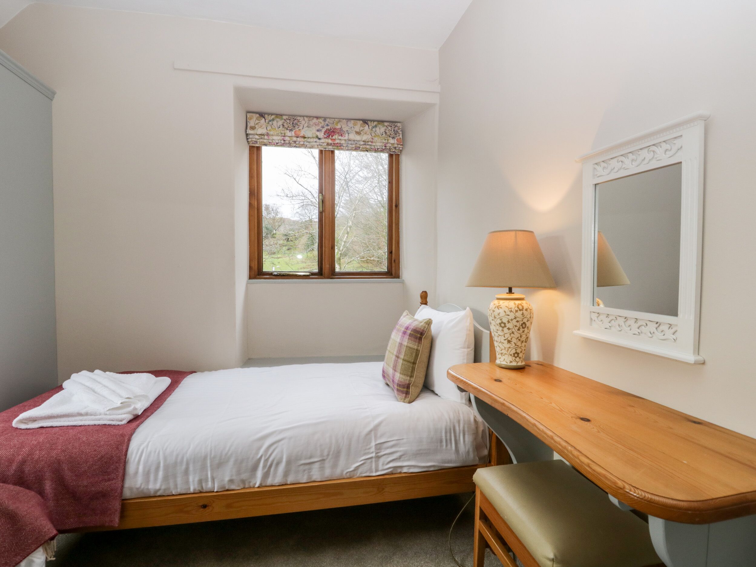 A bedroom with a single bed, a bedside table with a lamp, a mirror, and a window with a floral valance.