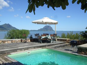 A poolside area with a wooden deck, outdoor seating, and a view of the sea and mountains.