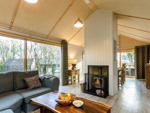 Una sala de estar acogedora con un sofá, una mesa de centro de madera con fruta, una chimenea y ventanas amplias que ofrecen vistas al exterior.