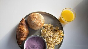 Un piatto con uova strapazzate, un croissant, una piccola ciotola di crema viola e un bicchiere di succo d'arancia.