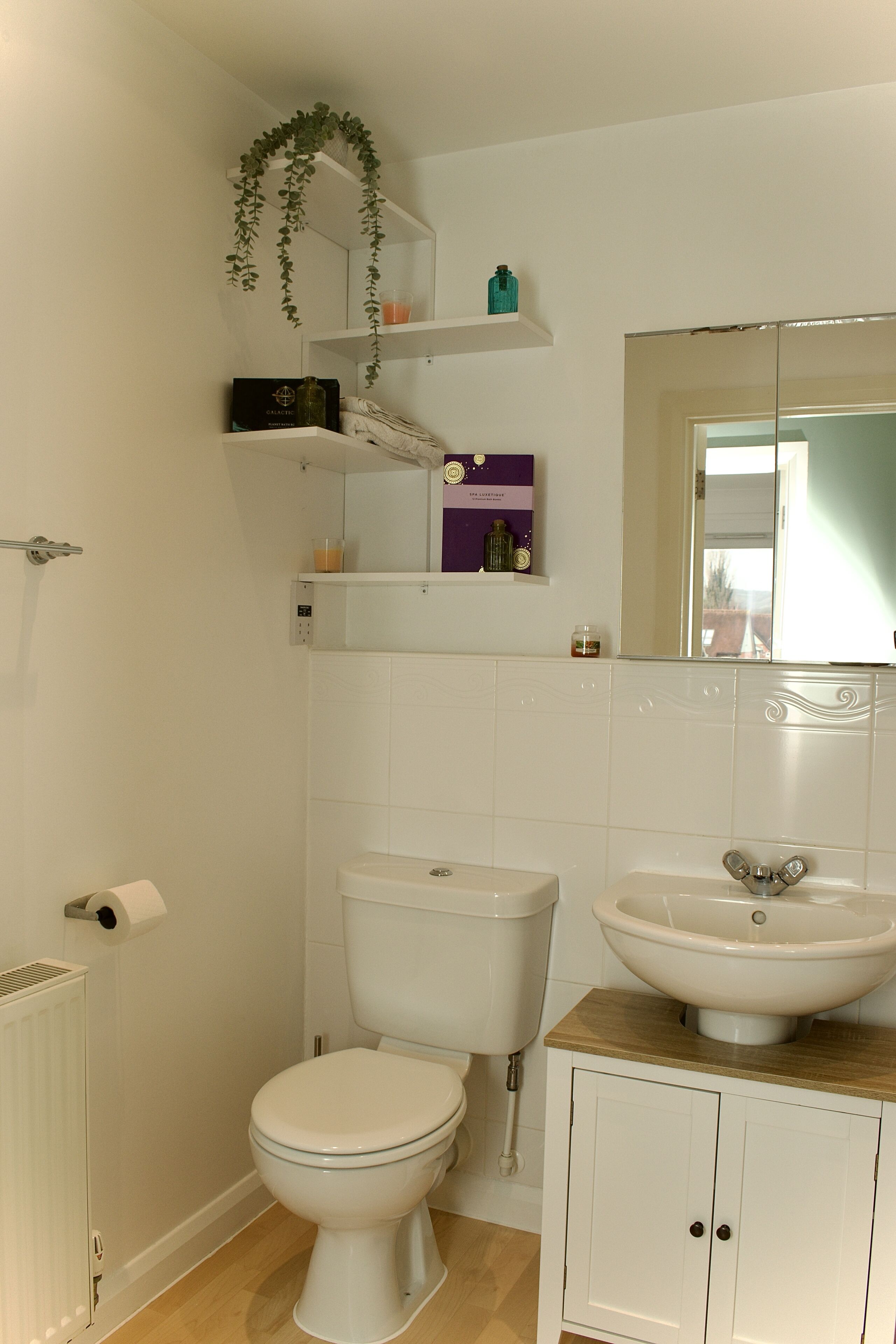 Un baño con inodoro, lavamanos y espejo. Hay repisas sobre el inodoro con diversos objetos.