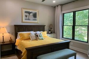 A bedroom with a large window, a bed with yellow bedding, a nightstand, and a bench.