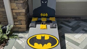 Batman-teemalla koristeltu penkki, jossa on pehmusteet ja logo. Penkki on sijoitettu kiviseinää ja ikkunaa vasten.