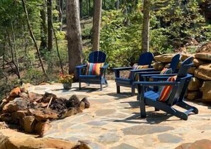 A stone patio with Adirondack chairs and a fire pit in a forested area.