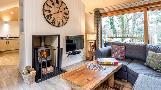 A living room with a large wall clock, a fireplace, a sofa, and a window with a view of trees. - Lodge 5 (Wareham)