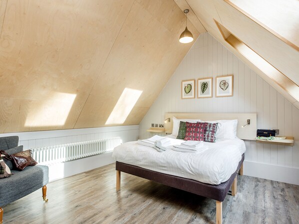 A bedroom with a sloped ceiling, a bed with white bedding, a grey sofa, and a radiator.