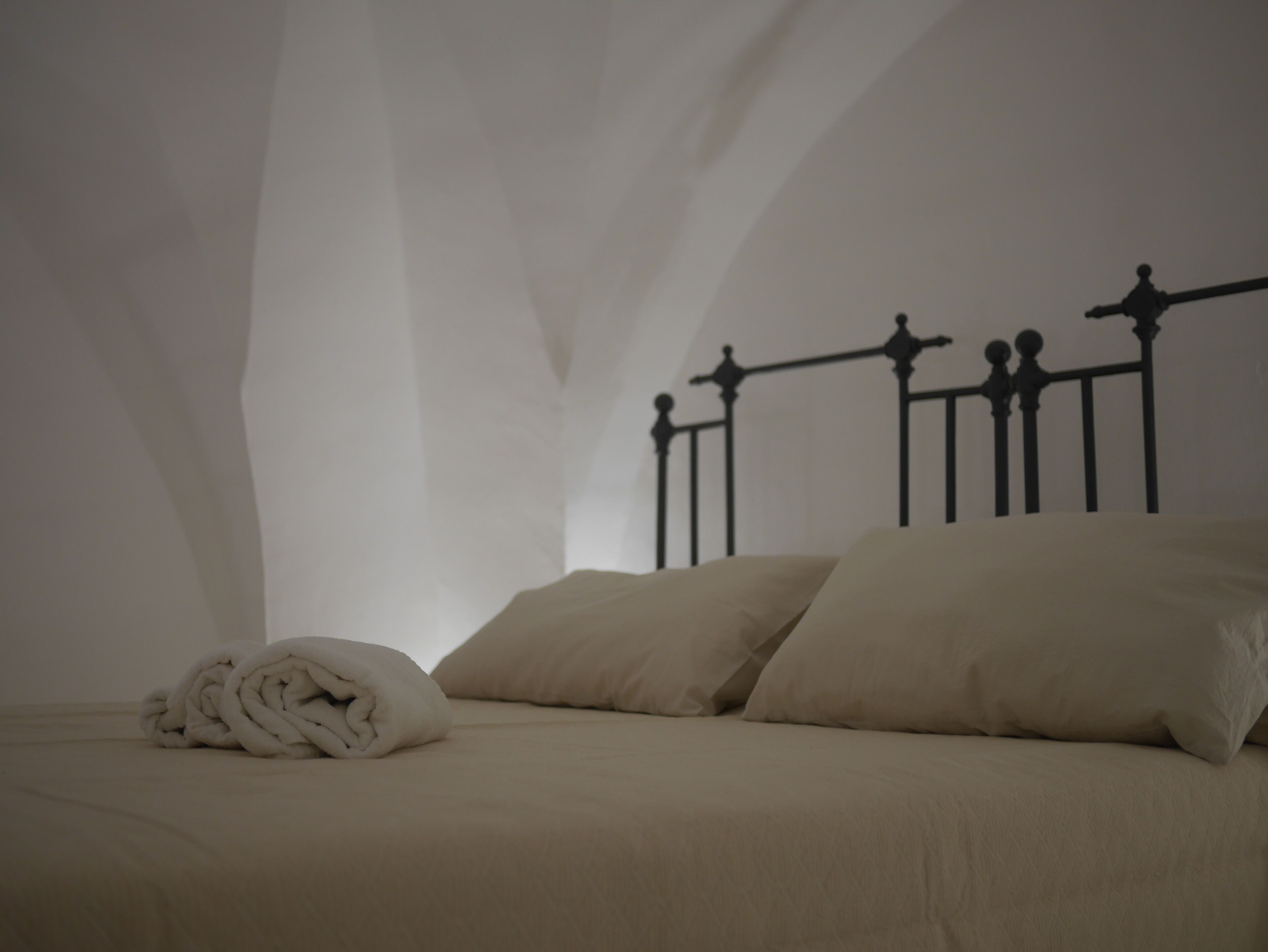 Una cama con cabecera de metal, dos almohadas, un edredón enrollado y una pared lisa.