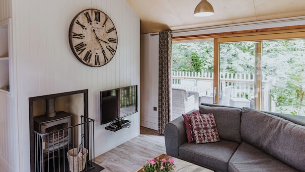 Un salón con un gran reloj de pared, chimenea, sofá y un amplio ventanal con vistas al verdor.