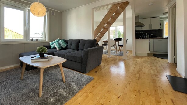 A modern living room with a grey sofa, a small wooden coffee table, and a grey rug. There is a wooden staircase leading to an upper level. The room has large windows and a kitchen area with white cabinets. - Flat With 2BR and Terrace Between Restaurants (Stavanger)