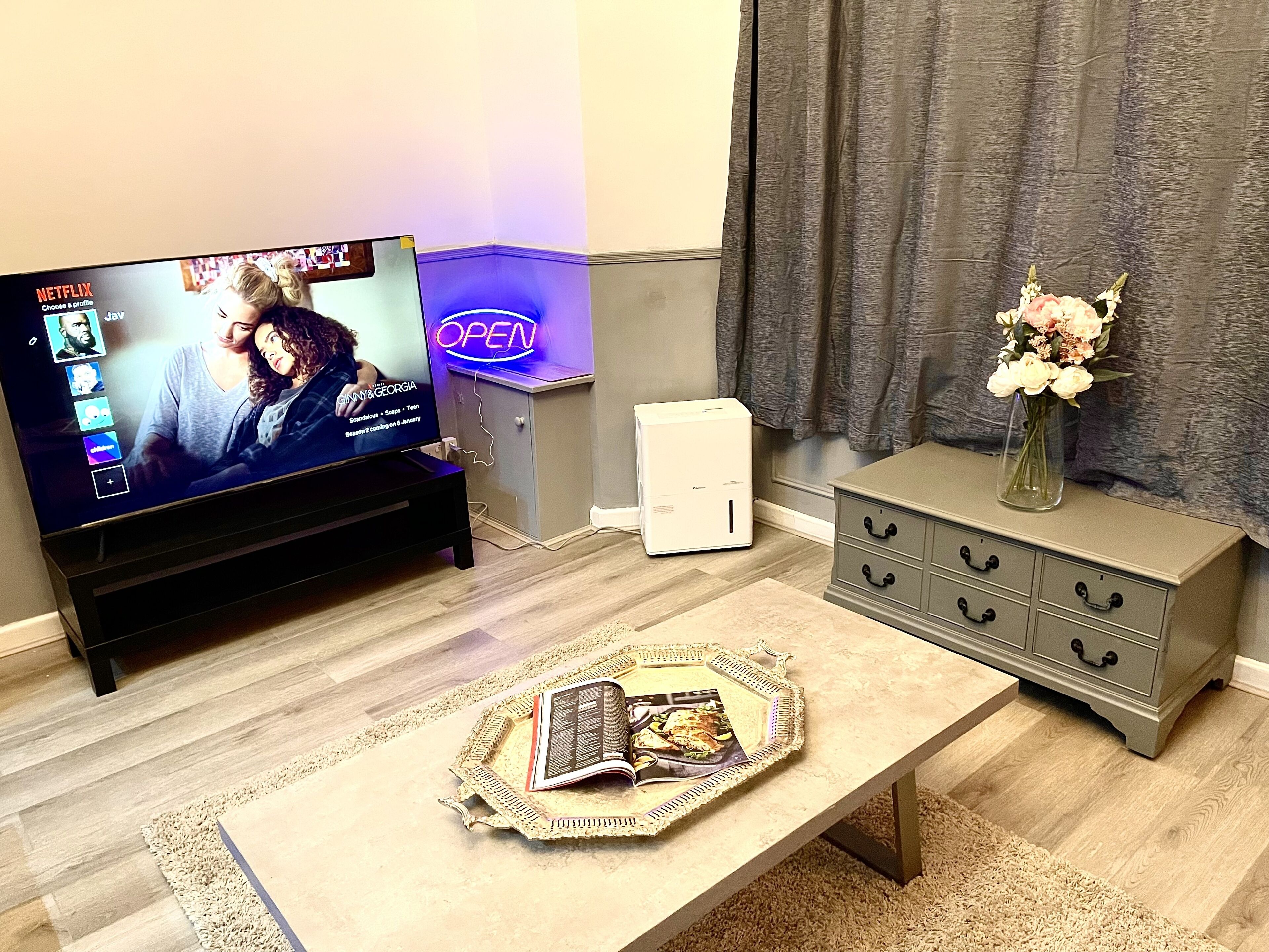 A living room with a flat-screen TV displaying a show, a coffee table with a book and a tray, a white heater, and a grey cabinet with a vase of flowers.