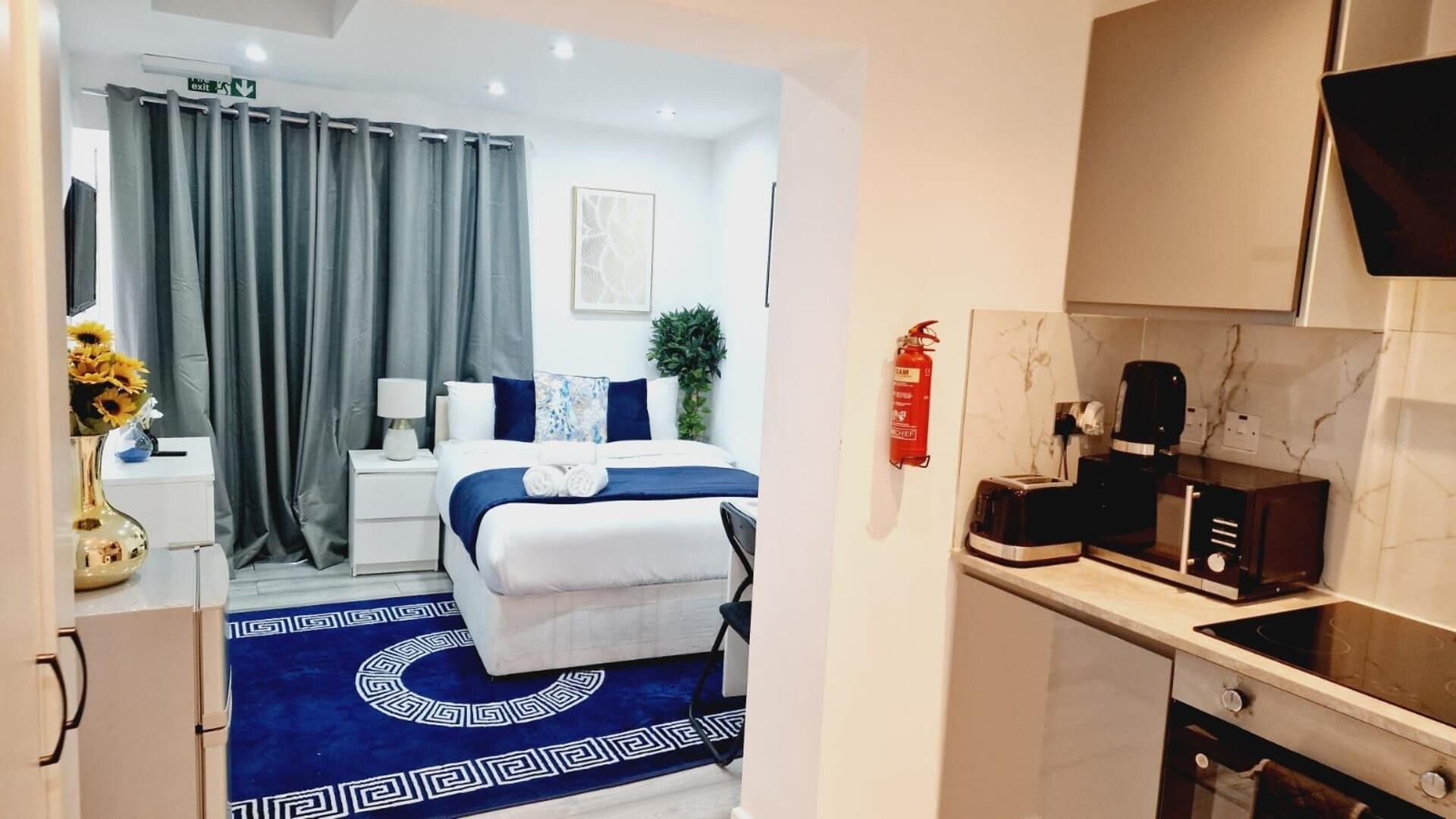 Une chambre d’hôtel comprenant un lit, une table de chevet, une lampe, un vase avec des fleurs, un tapis bleu à motifs, un coin cuisine avec une cuisinière, une machine à café et un extincteur.
