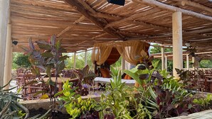 An outdoor seating area with wooden tables and chairs, covered by a wooden roof with a thatched ceiling.