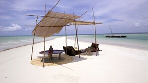 A beach scene with a thatched umbrella, a table, and chairs under the shade, a boat in the water, and a sandy beach.