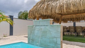 A swimming pool with a waterfall feature, surrounded by a thatched-roof structure and outdoor seating. - New-Modern Villa Island Escape, 10 Minute Drive to Beaches (Oranjestad)