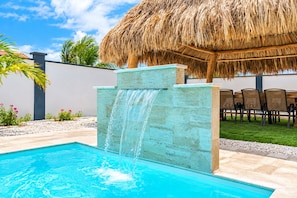 A swimming pool with a waterfall feature, surrounded by a thatched roof structure, outdoor seating, and a well-maintained garden. - New-Modern Villa Island Escape, 10 Minute Drive to Beaches (Oranjestad)