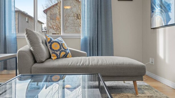 A modern living room with a grey sofa, a glass coffee table, and a view of a building through large windows. - Blue Moon | Playground | Backyard | Pet Friendly (Calgary)