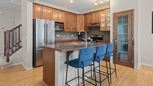 A modern kitchen with wooden cabinets, stainless steel appliances, and a bar area with blue barstools. - Blue Moon | Playground | Backyard | Pet Friendly (Calgary)