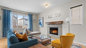 A modern living room with a fireplace, blue and yellow furniture, and a view of the outside. - Blue Moon | Playground | Backyard | Pet Friendly (Calgary)
