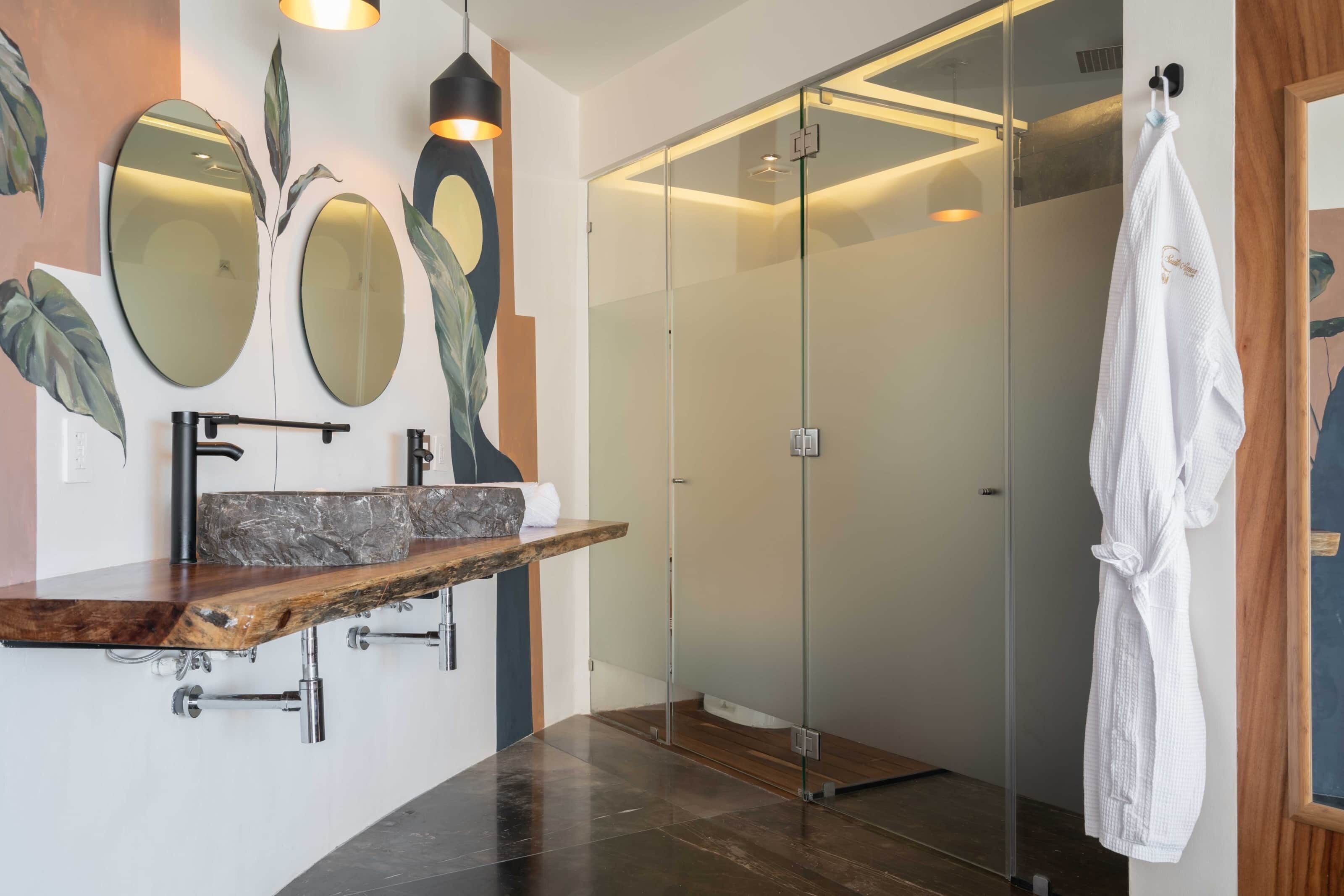 A modern bathroom with a glass-enclosed shower, a wooden vanity with a marble sink, and a white robe hanging on the wall.