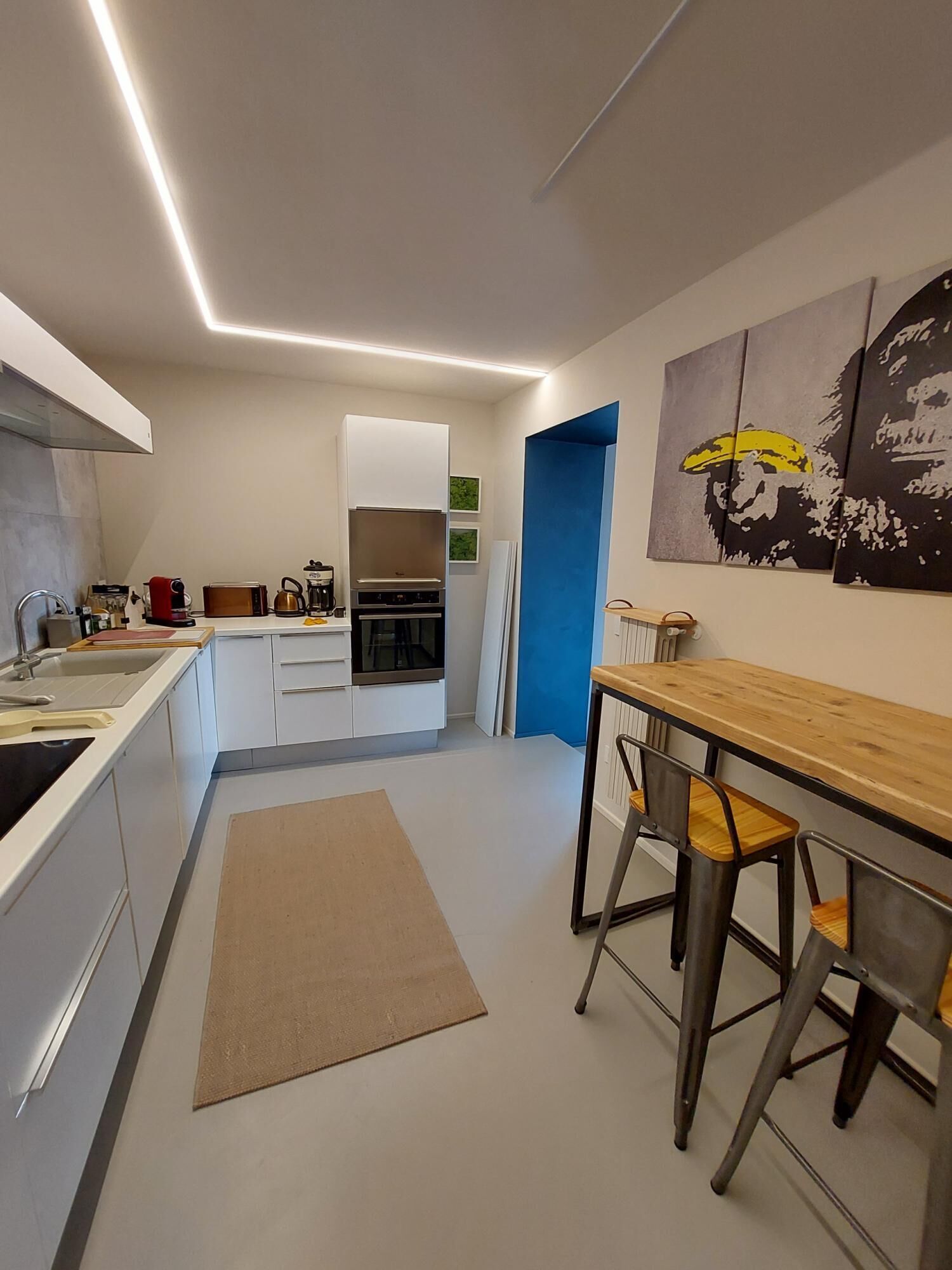 Une cuisine moderne avec des meubles blancs, une machine à café et un coin repas comprenant une table en bois et des chaises en métal.