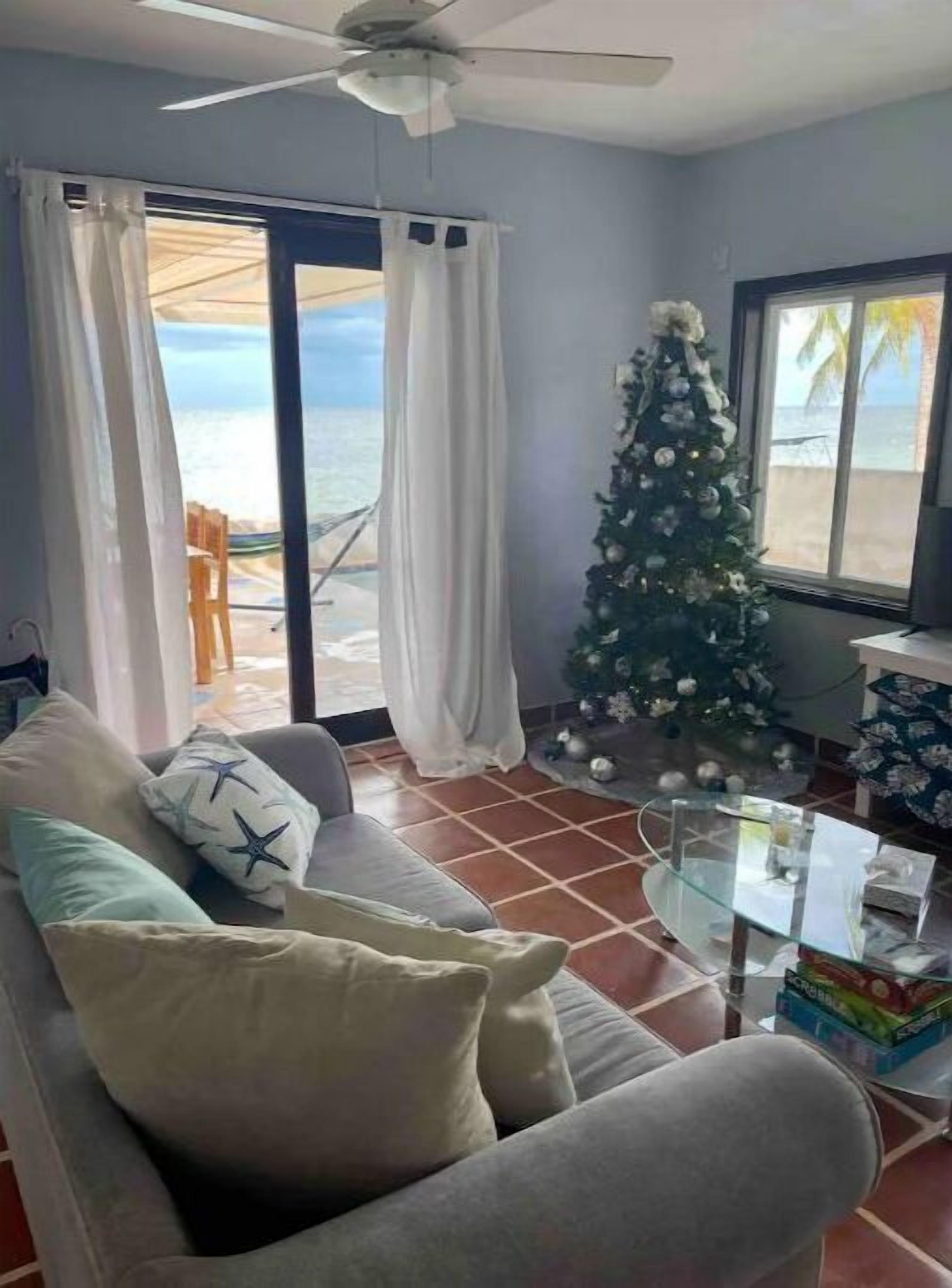 Een woonkamer met een bank, een glazen salontafel, een kerstboom en uitzicht op het strand door grote ramen.
