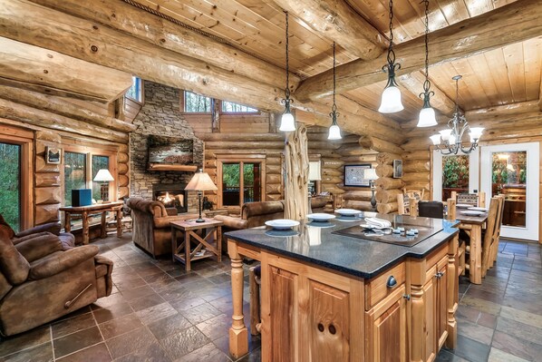 A log cabin interior with a stone fireplace, wooden furniture, and a large island with a black countertop.