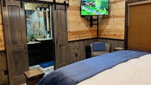 A bedroom with a bed, a TV mounted on the wall, a sliding door, and a small table. - Cozy Cabin W/ 2 King Suites, Pet Friendly, Hot tub (Broken Bow)