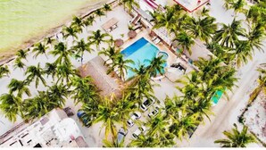 Vue aérienne d’un complexe hôtelier doté d’une piscine entourée de palmiers, d’une plage et de quelques bâtiments.