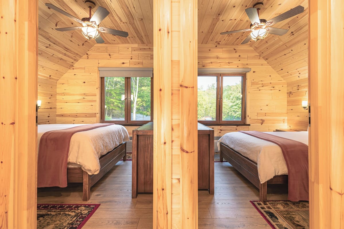 A cabin with two wooden beds, a ceiling fan, and a large window.