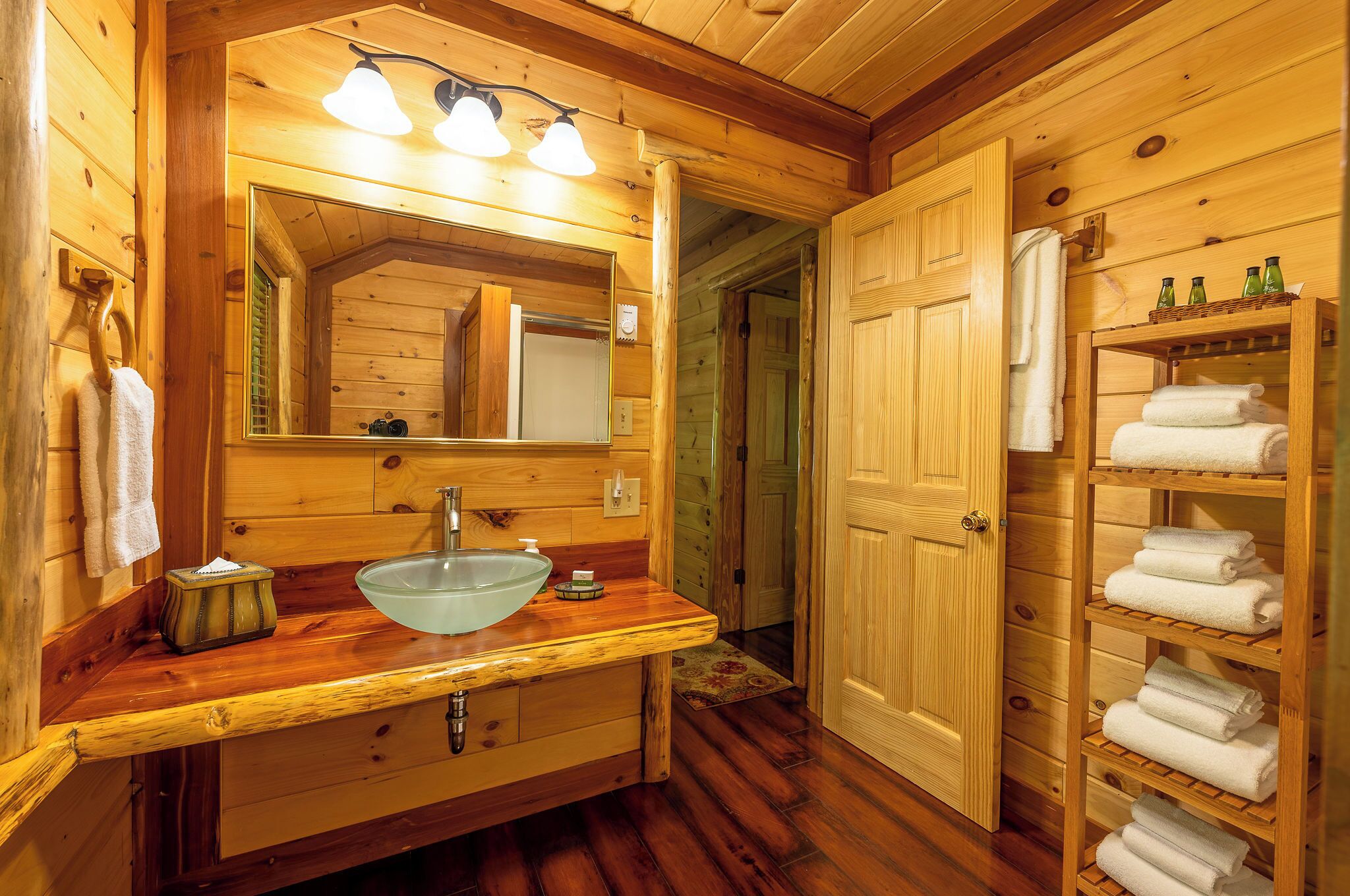 A wooden bathroom with a large mirror, a sink, and shelves with towels.