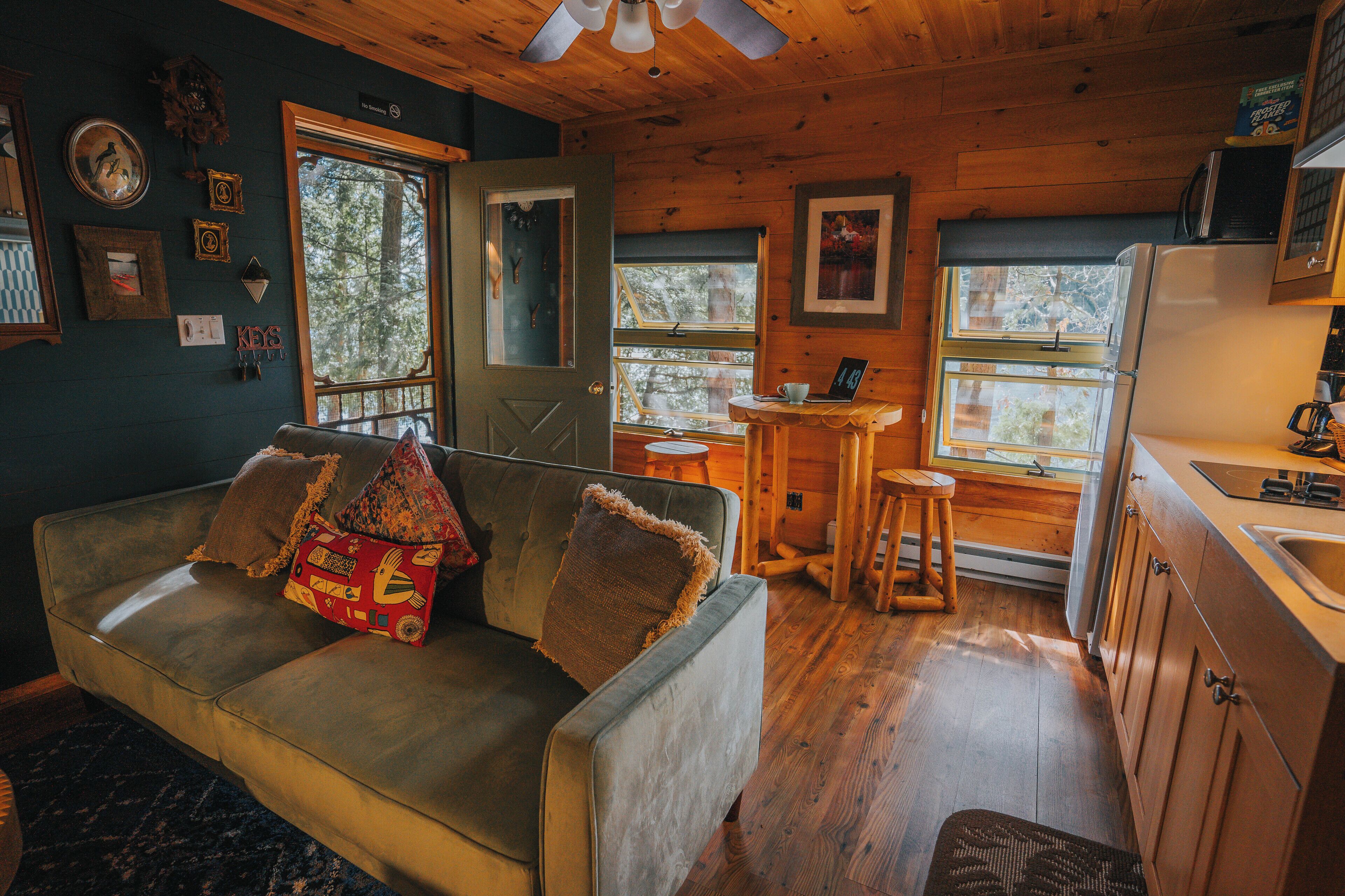 A cozy cabin interior with a sofa, wooden walls, a kitchen area, and large windows.