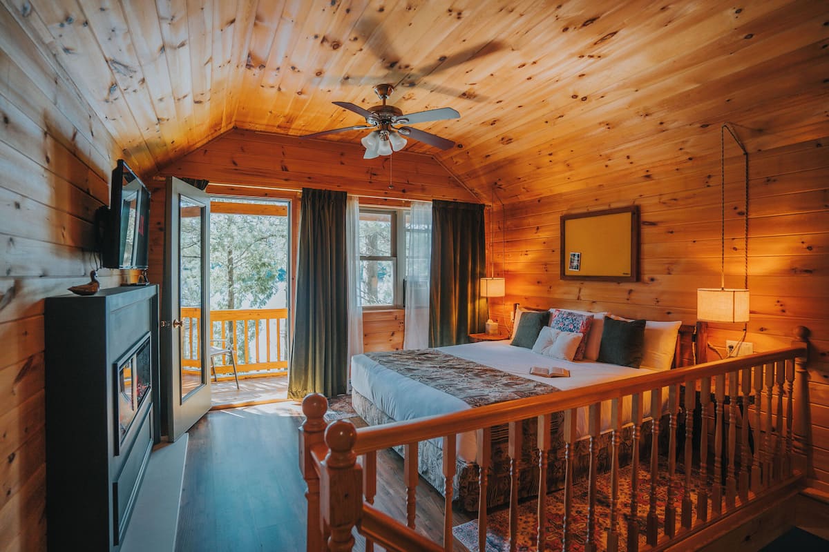 A cozy cabin bedroom with a bed, wooden walls, a ceiling fan, and a view of the outdoors.
