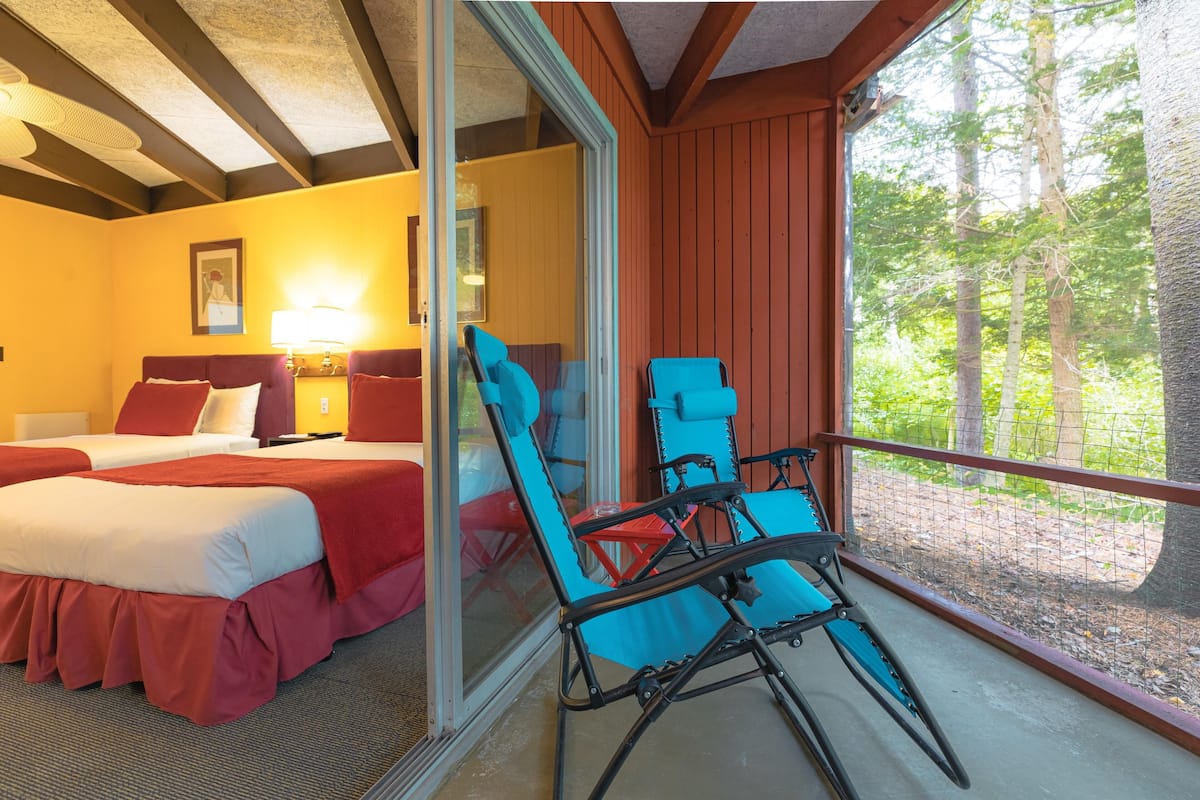 A room with two beds, a balcony with blue chairs, and a view of trees.