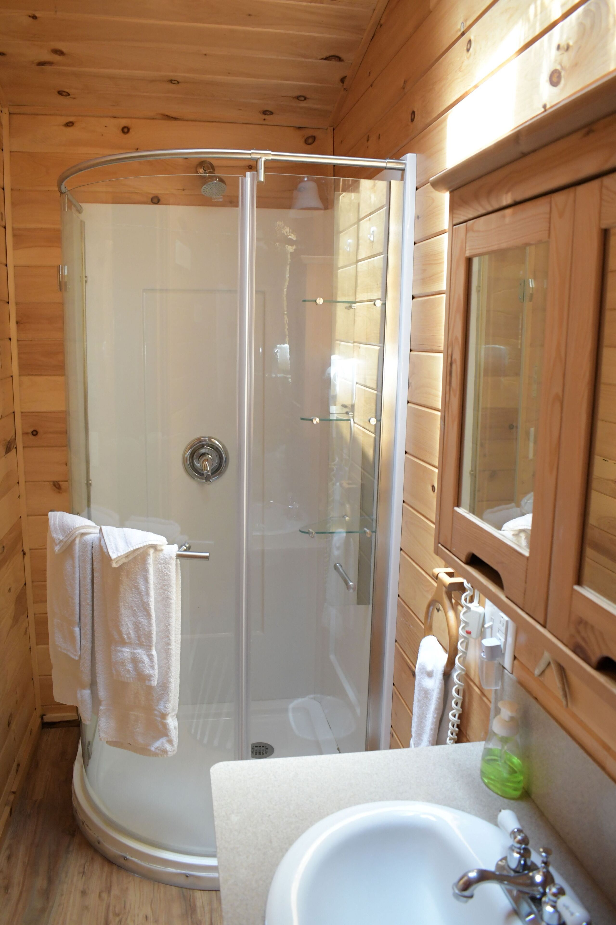 A wooden cabin bathroom with a glass-enclosed shower, a sink, and a mirror.