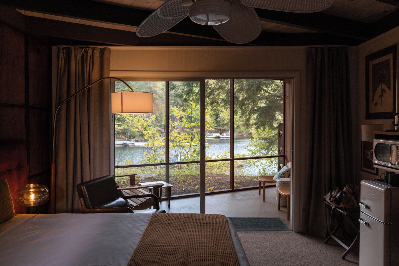 A hotel room with a bed, a desk, a chair, a dining table, a view of a lake, and a ceiling fan.