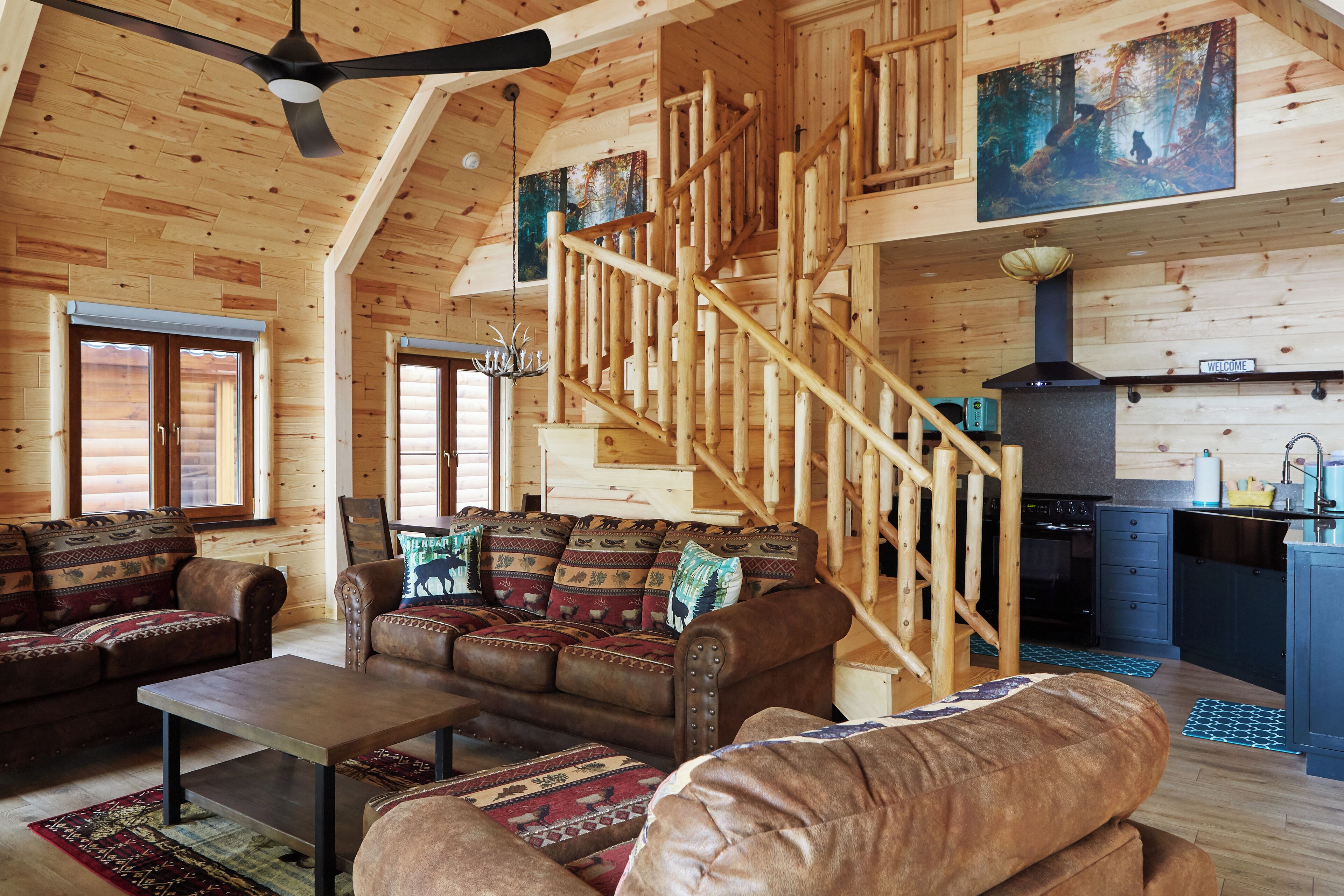 A log cabin interior with a wooden staircase, a living area with leather sofas, and a kitchen with a wood stove.