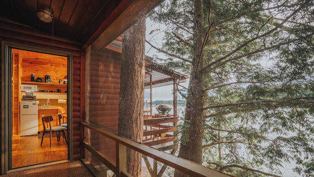 A cabin with a kitchen area visible through a glass door, a wooden deck, and a view of a forested area. - Motel Long Lake and Cottages (Long Lake)